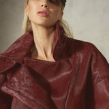 A close-up of a model wearing a textured, deep burgundy leather fur jacket with a high collar and an asymmetrical zipper, paired with a black baseball cap against a neutral gray background.