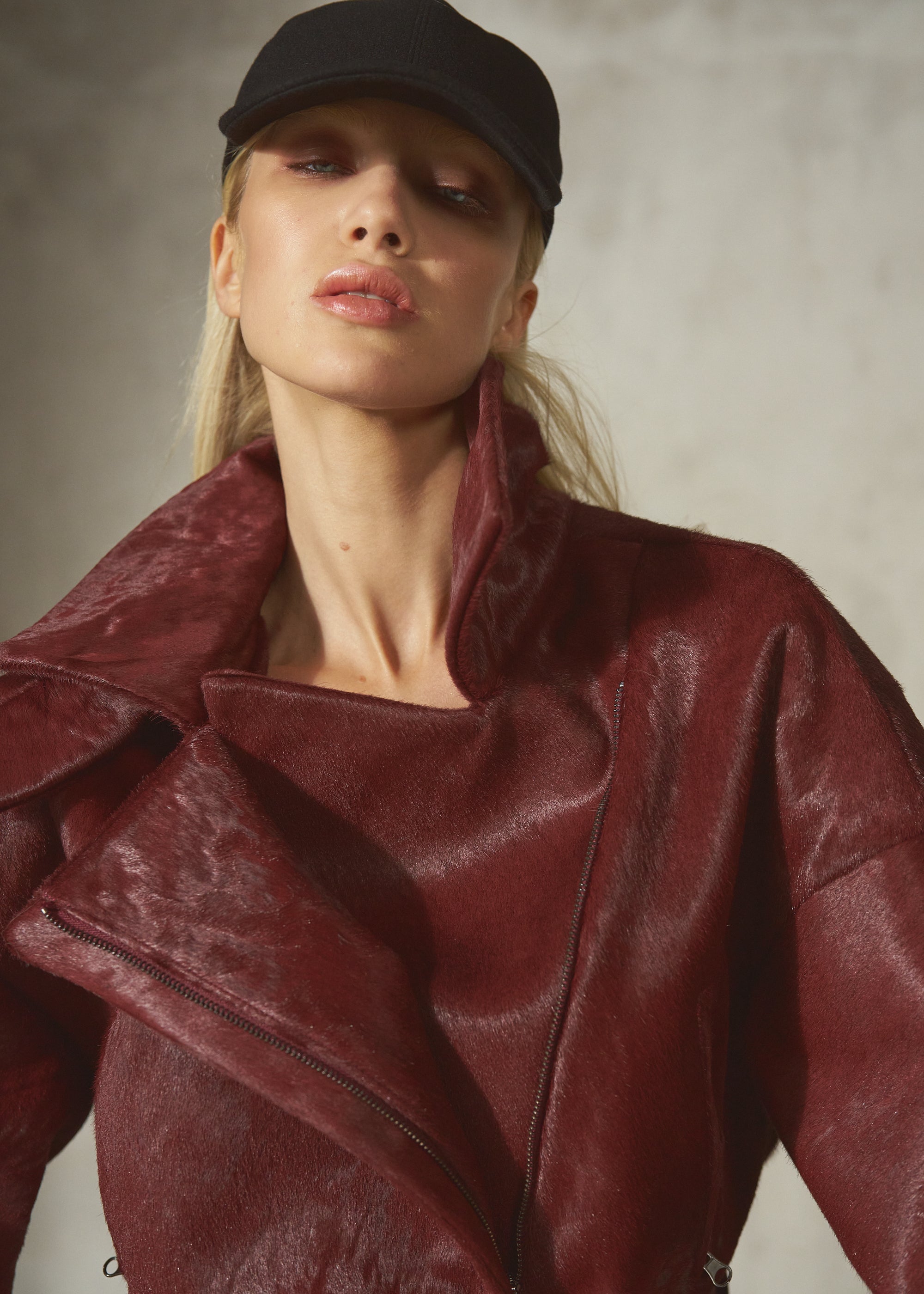 A close-up of a model wearing a textured, deep burgundy leather fur jacket with a high collar and an asymmetrical zipper, paired with a black baseball cap against a neutral gray background.