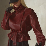 A model wearing a dark red ponyhair funnel neck jacket with a matching waist tie belt, black pants, black gloves, and a black baseball cap, posed against a textured gray wall.