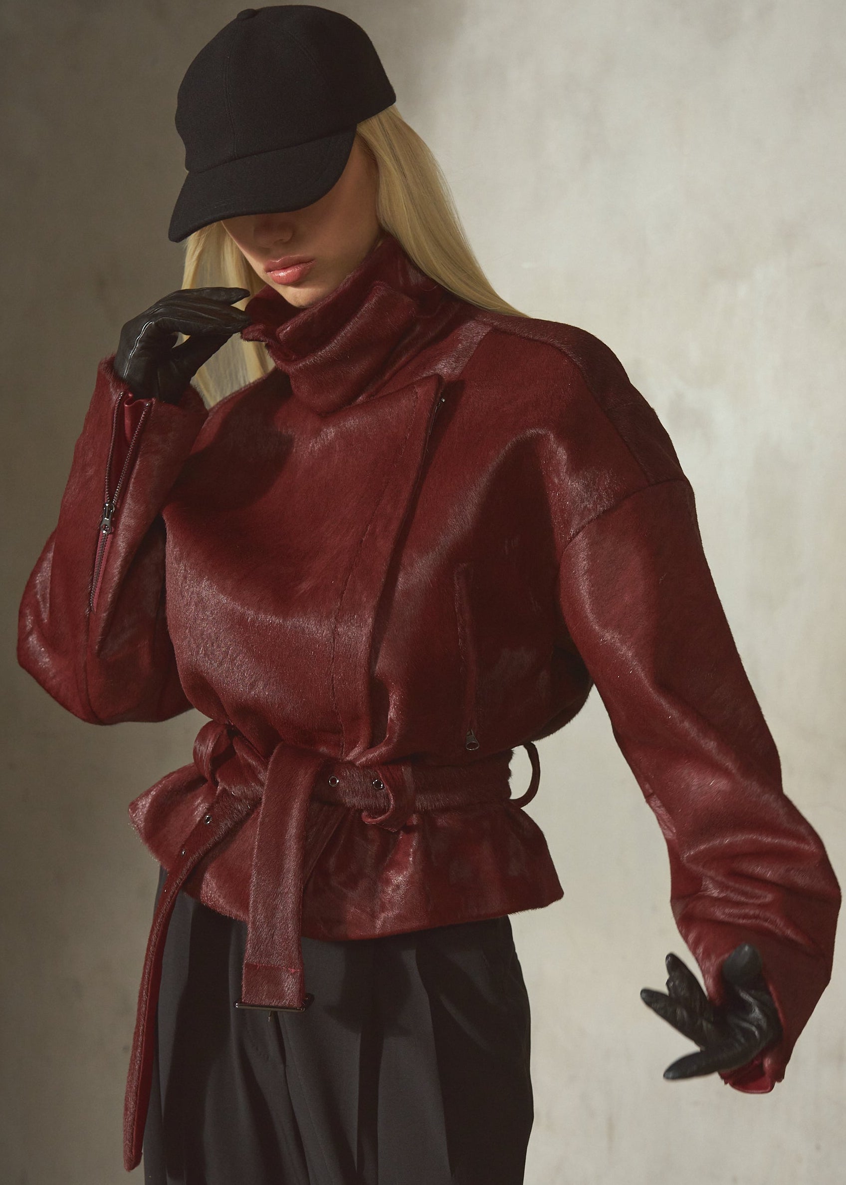 A model wearing a dark red ponyhair funnel neck jacket with a matching waist tie belt, black pants, black gloves, and a black baseball cap, posed against a textured gray wall.