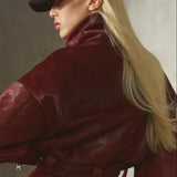 A woman wearing a deep burgundy fur jacket with a matching belt, black pants, a black baseball cap, and black gloves, photographed against a textured concrete wall.