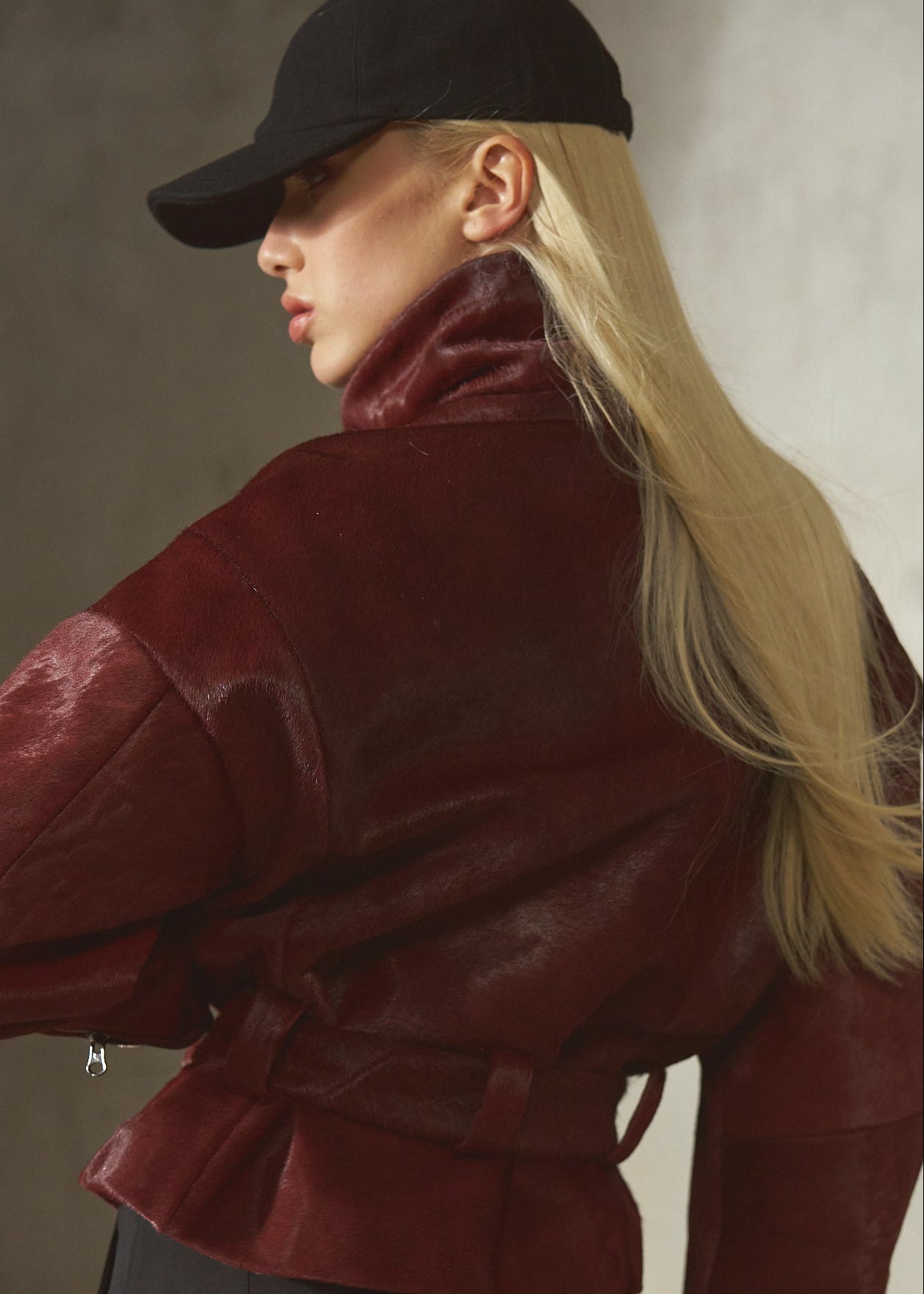 A woman wearing a deep burgundy fur jacket with a matching belt, black pants, a black baseball cap, and black gloves, photographed against a textured concrete wall.