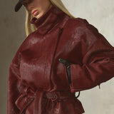 A model wearing a deep burgundy ponyhair jacket with a high collar and matching tie belt, styled with a black baseball cap and black pants against a textured gray wall.