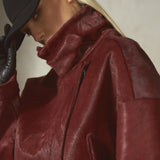 A close-up of a model wearing a textured, deep burgundy fur jacket with a high collar, black gloves, and a black baseball cap against a neutral gray background.