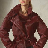 A model wearing a dark burgundy ponyhair jacket with a matching belt, black pants, and a black baseball cap, posing with a textured gray wall.
