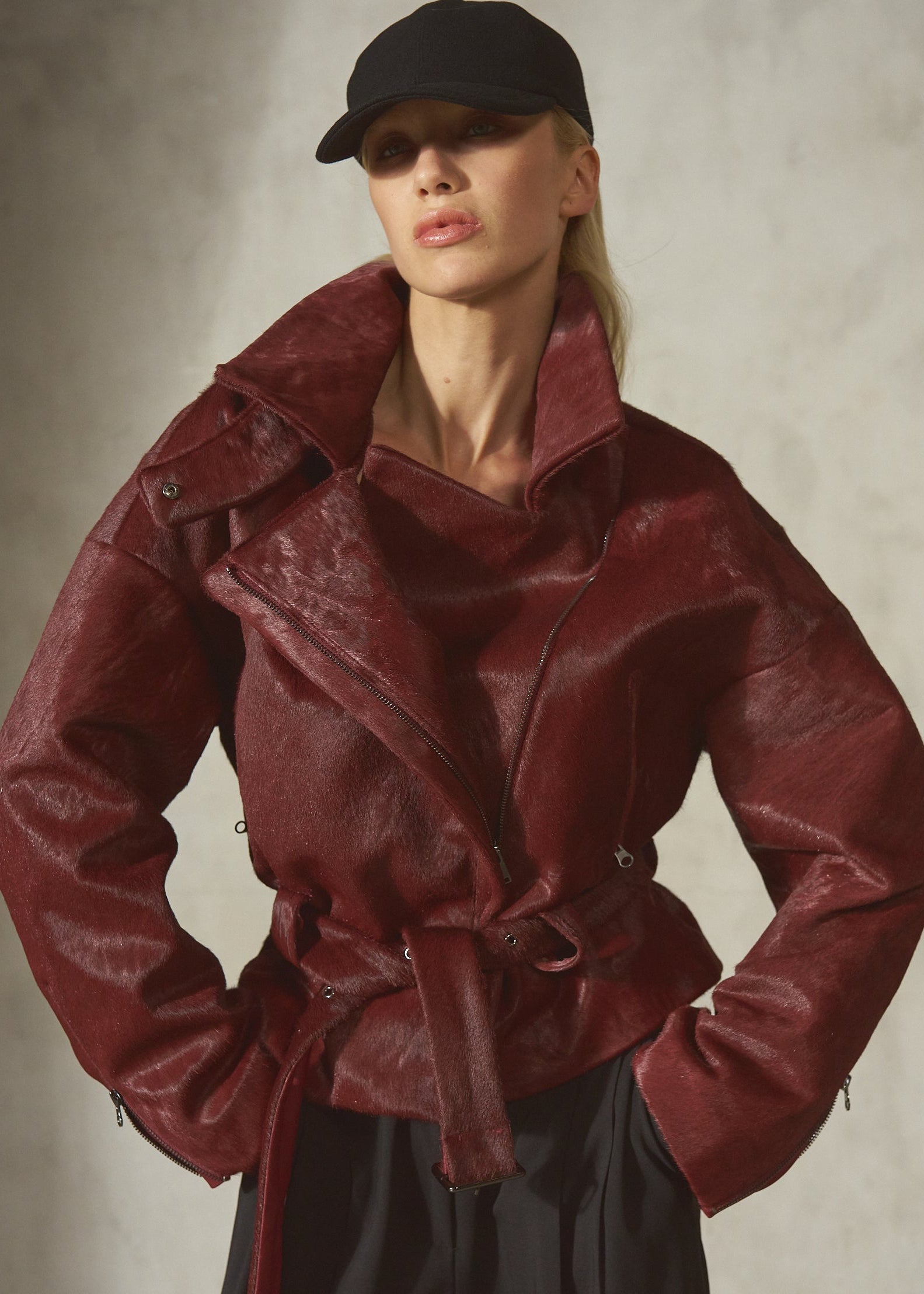 A model wearing a dark burgundy ponyhair jacket with a matching belt, black pants, and a black baseball cap, posing with a textured gray wall.