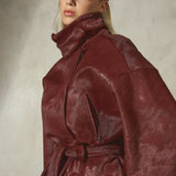 A model wearing an oversized fur burgundy jacket with a high collar and belt, paired with black pants and a black baseball cap, set against a textured gray studio background.
