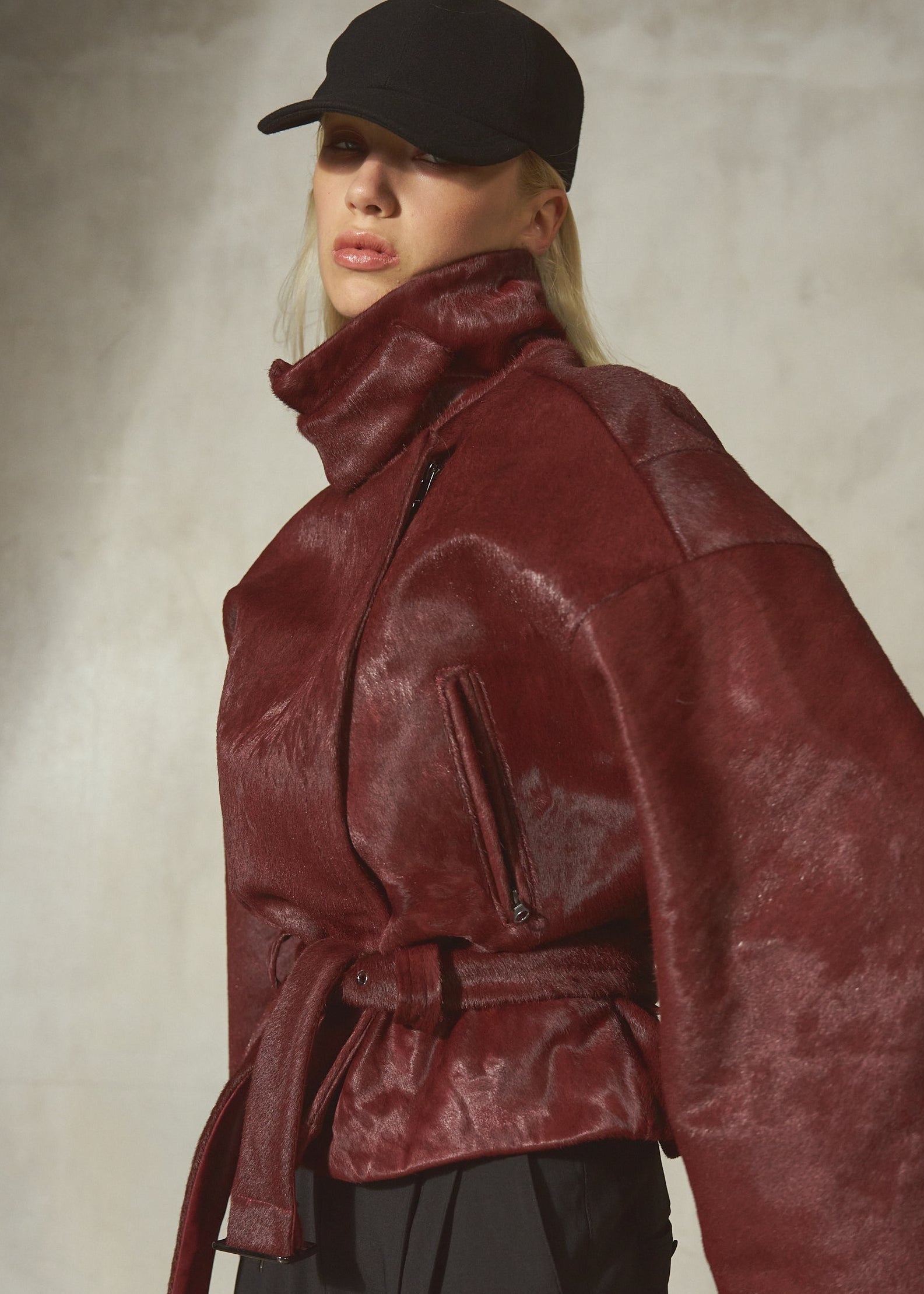 A model wearing an oversized fur burgundy jacket with a high collar and belt, paired with black pants and a black baseball cap, set against a textured gray studio background.