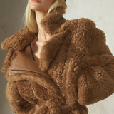 A blonde model wearing a voluminous brown teddy fur jacket with a matching belt, a black baseball cap, and black pants, posing against a textured gray wall.