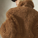 A model shown from the back wearing a brown teddy-fur cropped jacket, a black baseball cap, and dark pants against a textured gray wall.