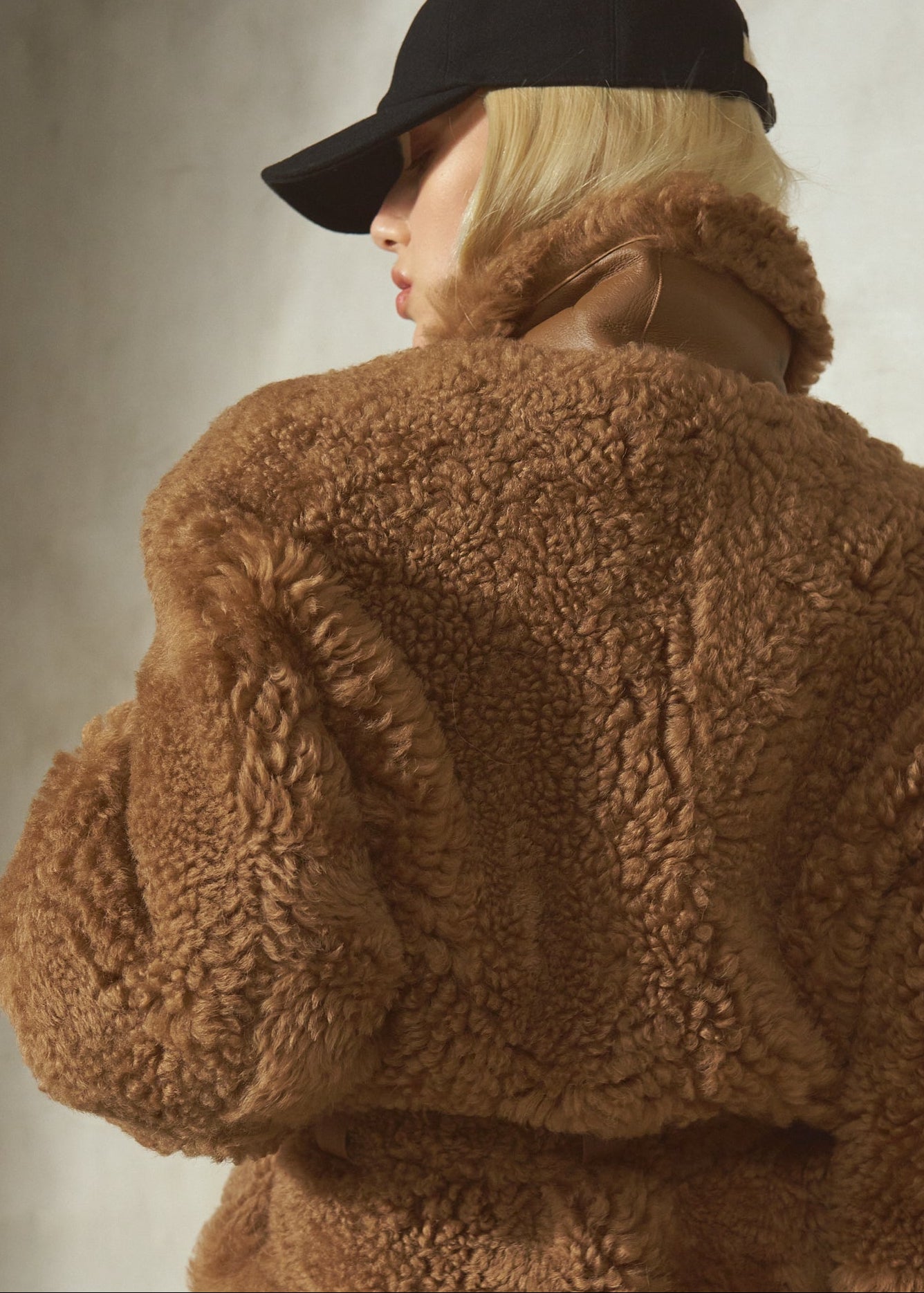 A model shown from the back wearing a brown teddy-fur cropped jacket, a black baseball cap, and dark pants against a textured gray wall.