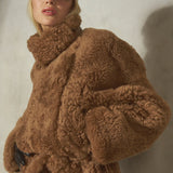 A model wearing a voluminous, high-collar brown curly shearling jacket with a matching tie belt, styled with a black cap and black pants against a textured gray wall.