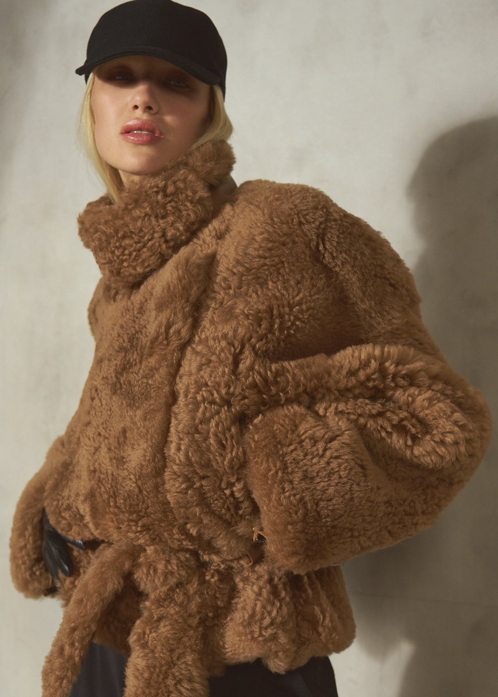 A model wearing a voluminous, high-collar brown curly shearling jacket with a matching tie belt, styled with a black cap and black pants against a textured gray wall.