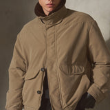 A model wearing a tan utility-style puffer jacket with a high shearling collar, black cap, and black gloves, standing against a textured gray wall.
