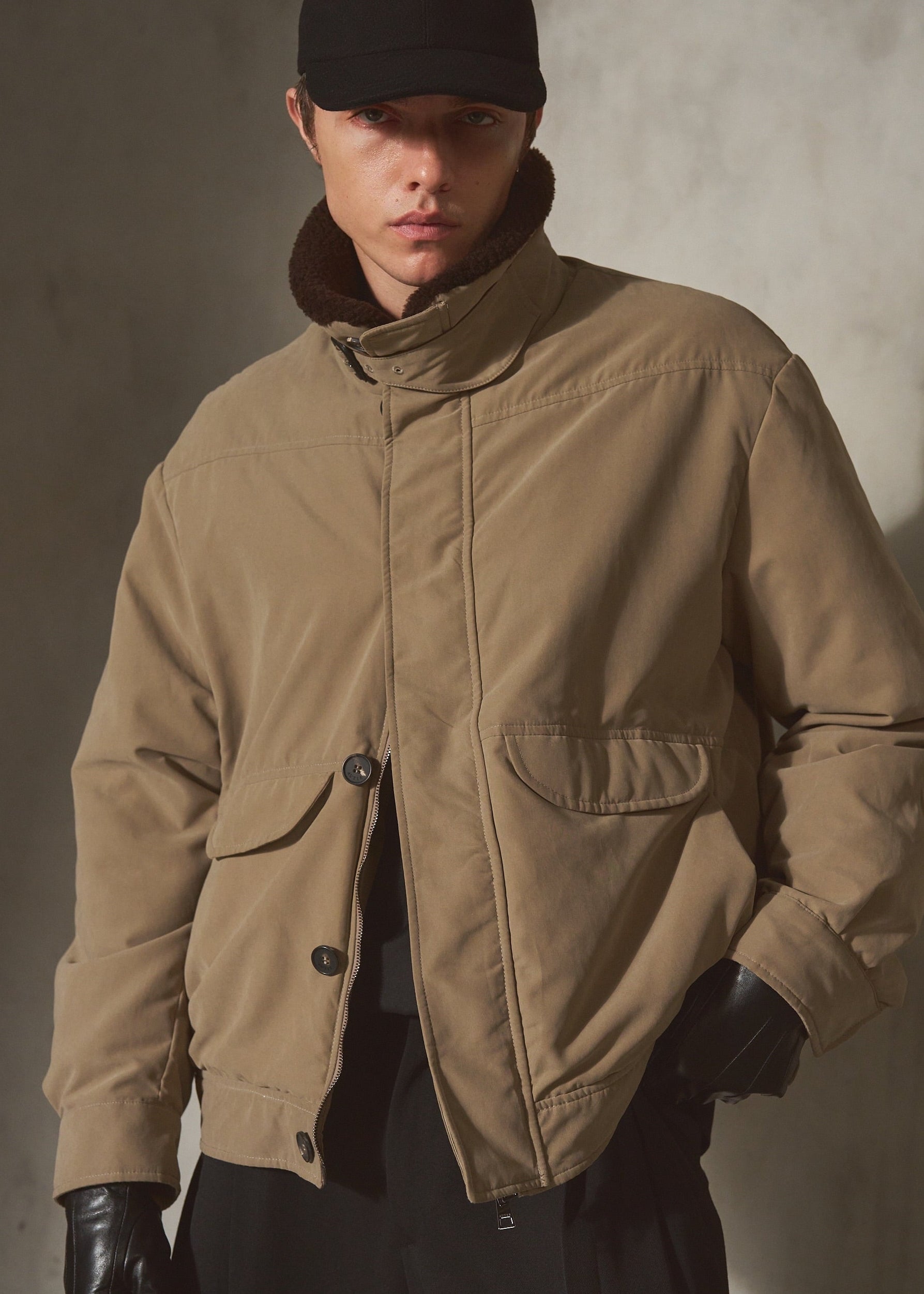 A model wearing a tan utility-style puffer jacket with a high shearling collar, black cap, and black gloves, standing against a textured gray wall.