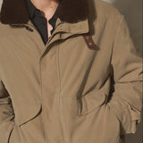A man wearing a beige utility jacket with a dark brown shearling collar, a black baseball cap, and a black shirt, standing against a textured gray concrete wall.