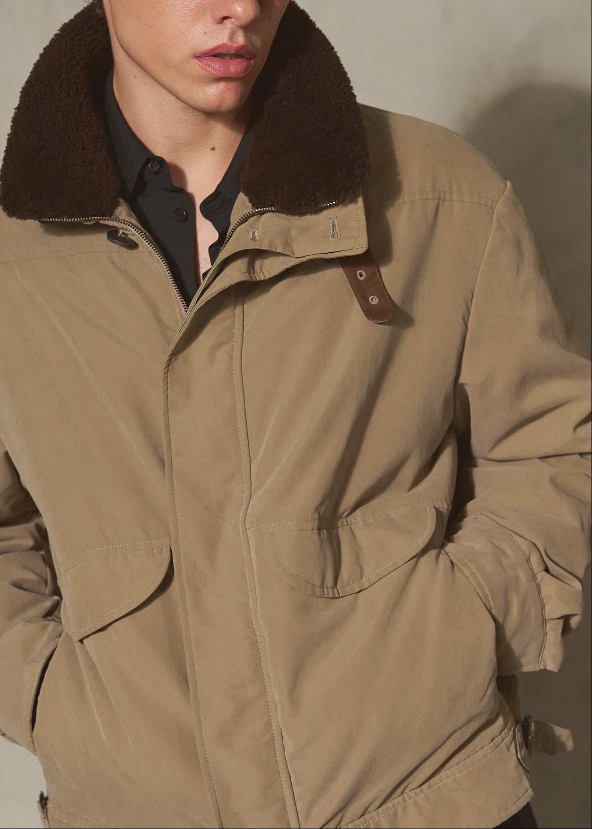 A man wearing a beige utility jacket with a dark brown shearling collar, a black baseball cap, and a black shirt, standing against a textured gray concrete wall.