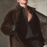 A man wearing a dark brown plush shealring jacket over a black shirt and trousers, accessorized with a black cap, black gloves, and a black leather coat draped off his shoulder, set against a textured gray studio background.