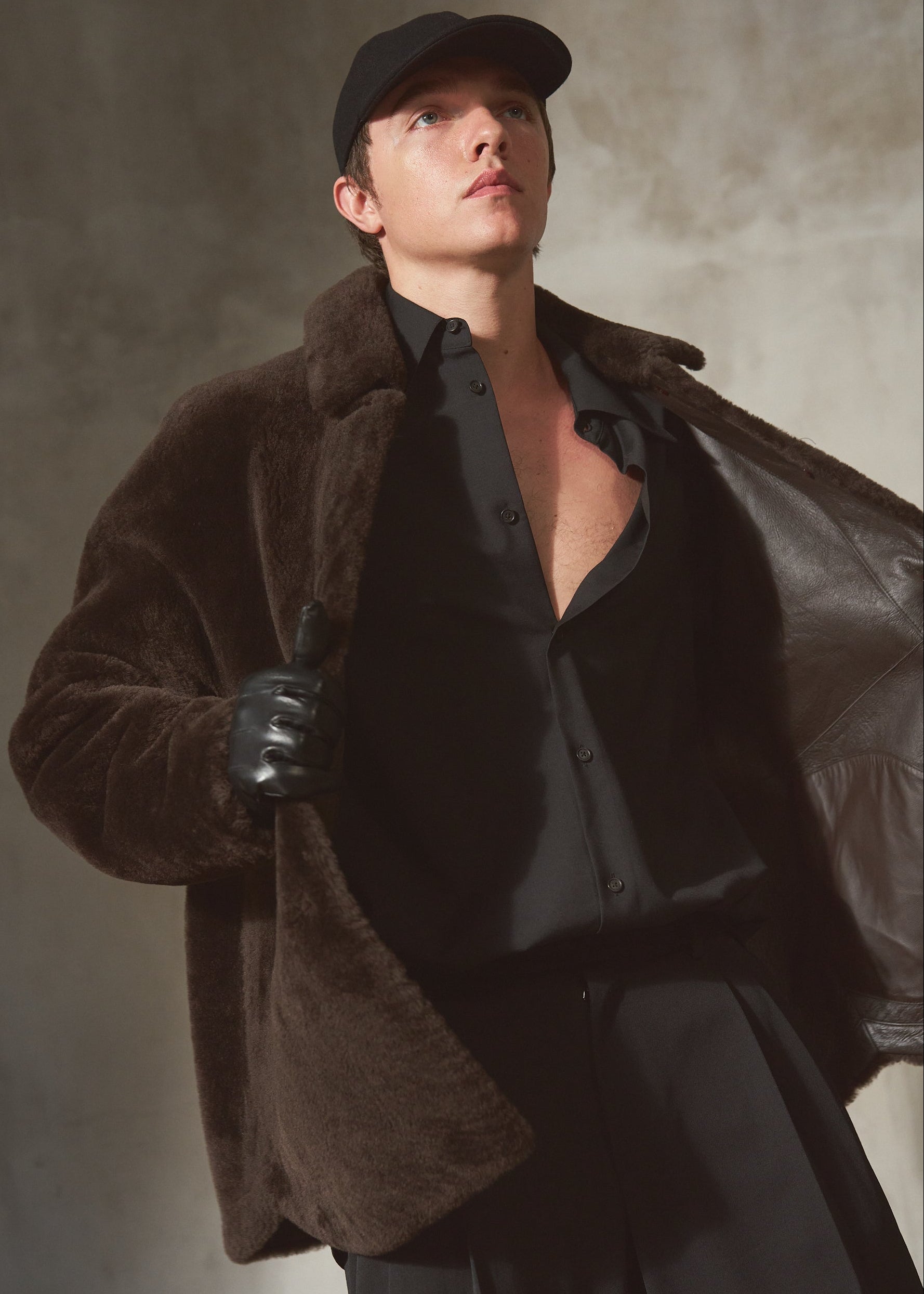 A man wearing a dark brown plush shealring jacket over a black shirt and trousers, accessorized with a black cap, black gloves, and a black leather coat draped off his shoulder, set against a textured gray studio background.
