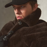 A close-up portrait of a person wearing a black baseball cap, a dark brown fur-trimmed coat, and black gloves against a neutral gray background.