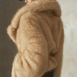 A model wearing a voluminous natural-colored faux fur coat with a large collar, paired with a black baseball cap and black trousers, standing against a textured concrete wall.