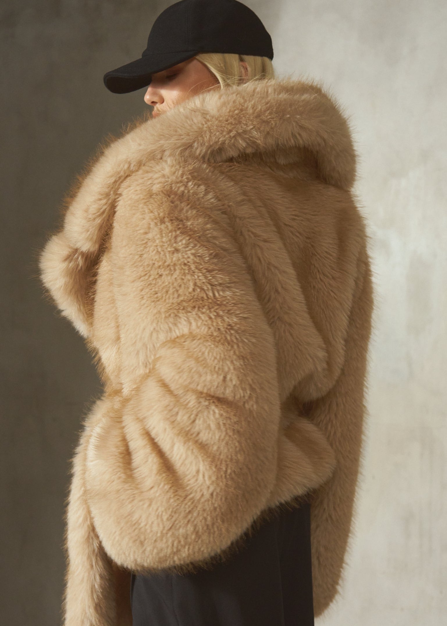 A model wearing a voluminous natural-colored faux fur coat with a large collar, paired with a black baseball cap and black trousers, standing against a textured concrete wall.
