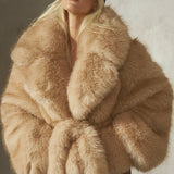 A model wearing a voluminous, oversized camel-colored faux fur jacket with a matching tie belt, styled with a black cap and black trousers, standing against a textured concrete wall.