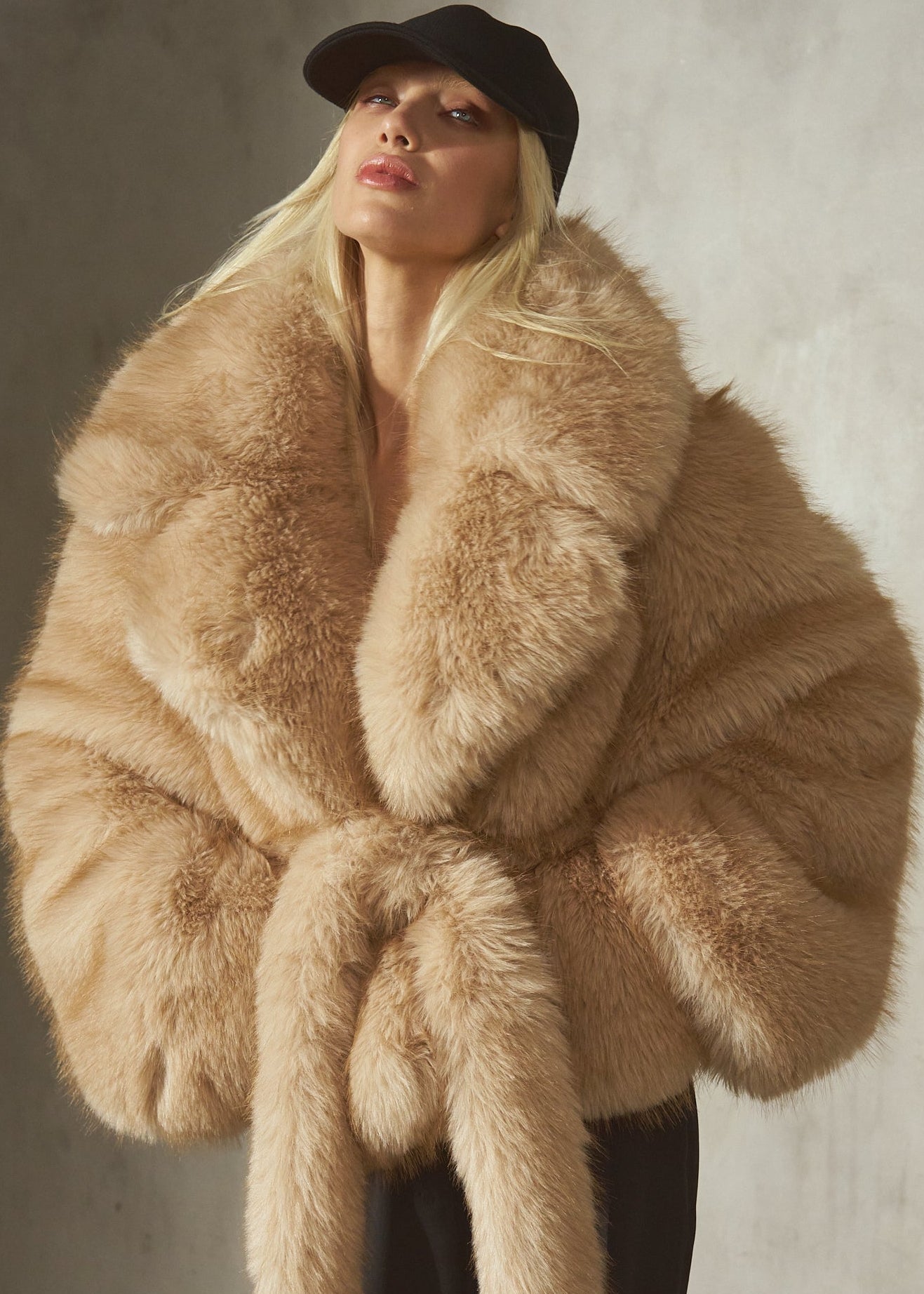 A model wearing a voluminous, oversized camel-colored faux fur jacket with a matching tie belt, styled with a black cap and black trousers, standing against a textured concrete wall.