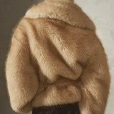 A model shown from the back wearing a voluminous tan faux fur jacket with a large matching collar, a black cap, and dark bottoms, standing in front of a textured gray wall.