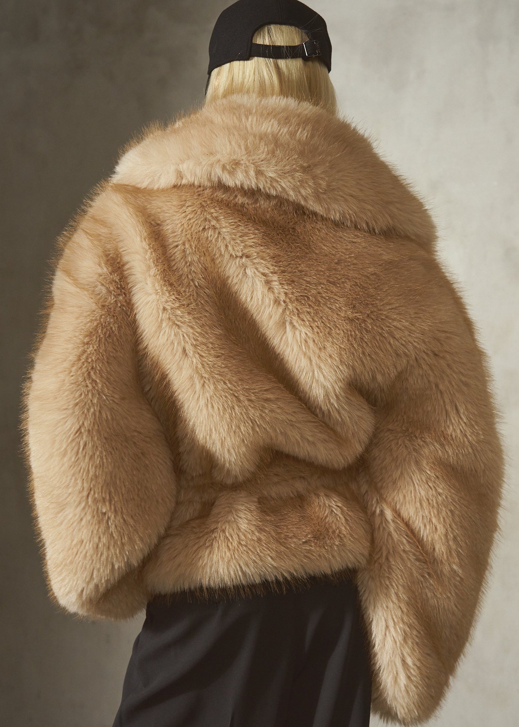 A model shown from the back wearing a voluminous tan faux fur jacket with a large matching collar, a black cap, and dark bottoms, standing in front of a textured gray wall.