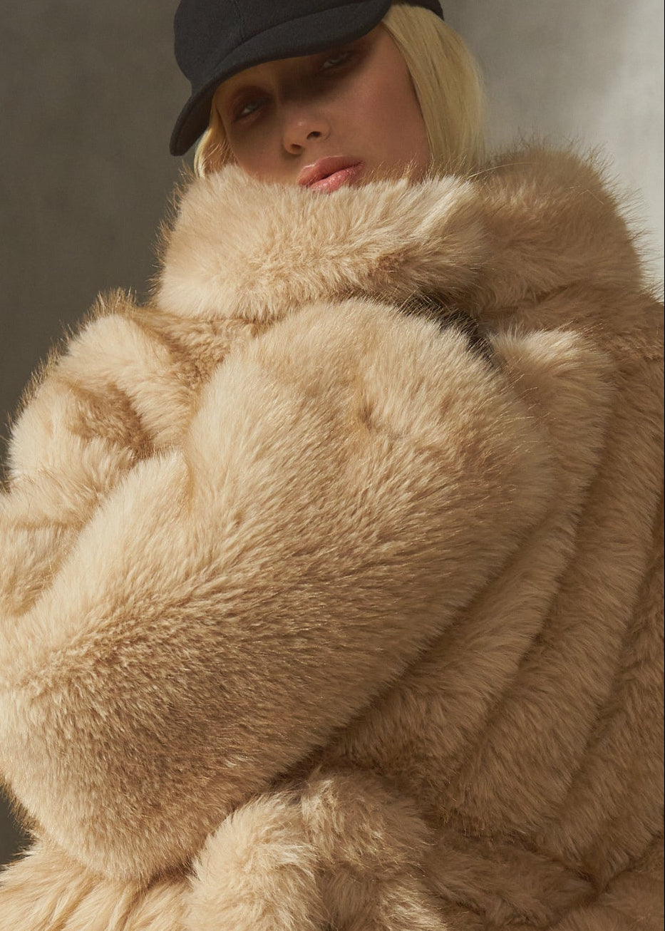 A model wearing a voluminous, oversized camel-colored faux fur coat with a high collar, belted, paired with black pants and a black cap, standing in front of a textured concrete wall.
