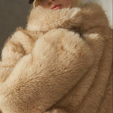 A model wearing a voluminous, oversized camel-colored faux fur coat with a high collar, belted, paired with black pants and a black cap, standing in front of a textured concrete wall.