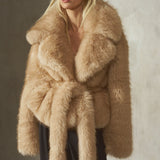 A model wearing a voluminous beige faux fur wrap coat with a matching belt, black pants, and a black baseball cap, standing in front of a textured concrete wall.