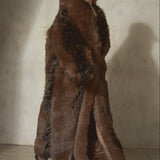 A model wearing a long, luxurious dark brown faux-fur coat with black gloves, sheer black tights, and black high heels, standing against a textured concrete wall.