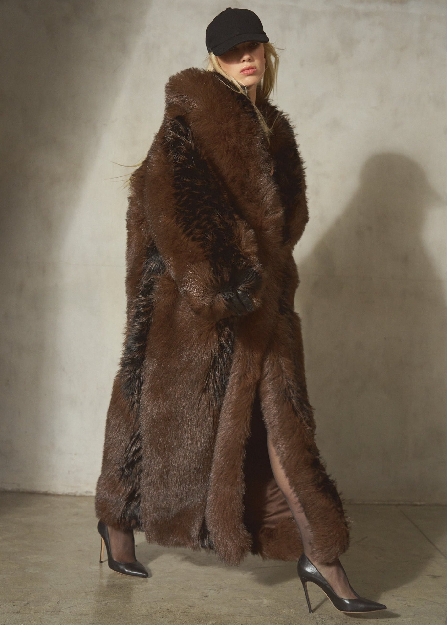 A model wearing a long, luxurious dark brown faux-fur coat with black gloves, sheer black tights, and black high heels, standing against a textured concrete wall.