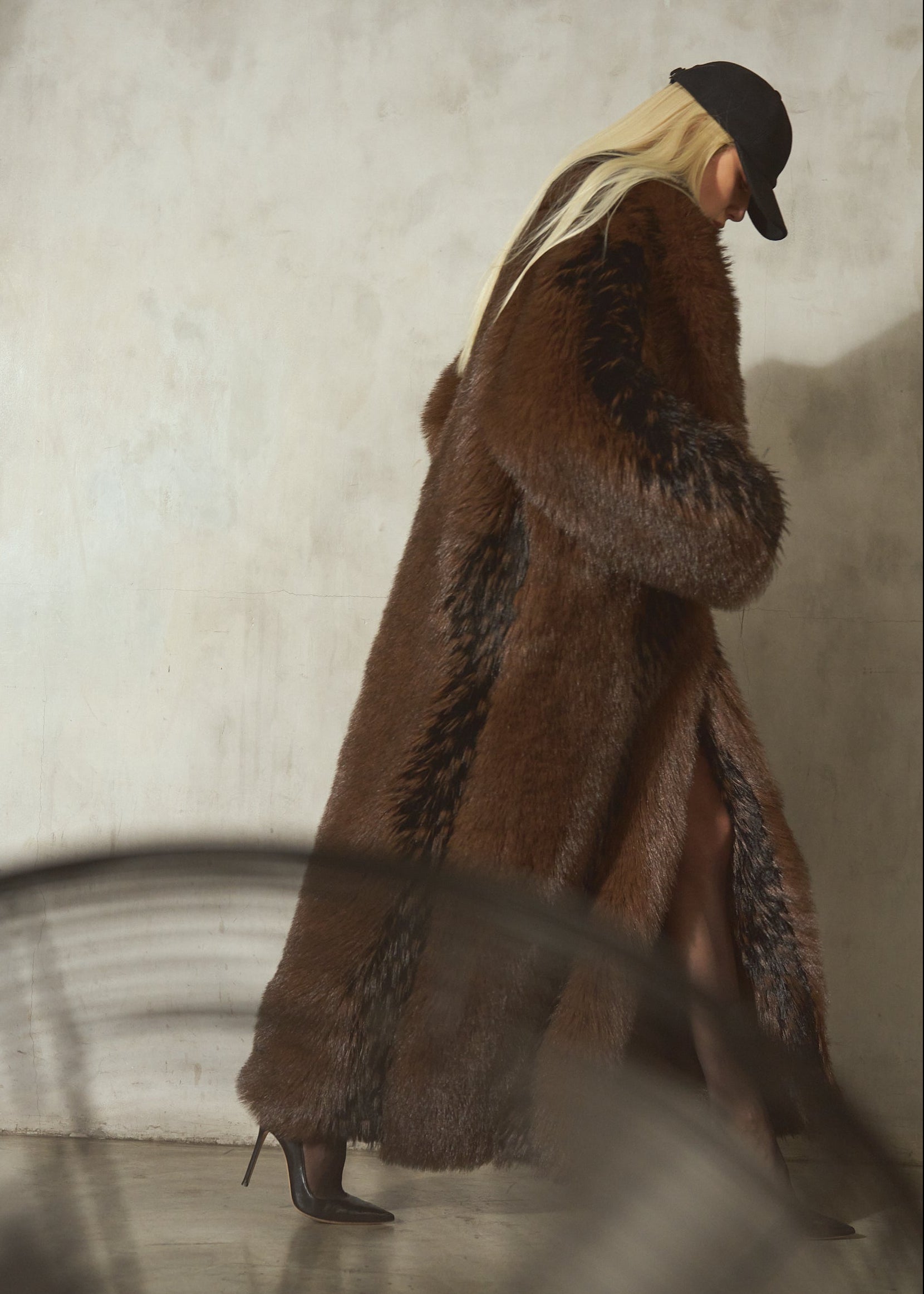 A woman wearing a long brown and black faux fur coat, styled with a black wool cap and black high heels.