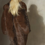 A blonde model viewed from behind wearing a voluminous dark brown and black faux fur floor length coat.