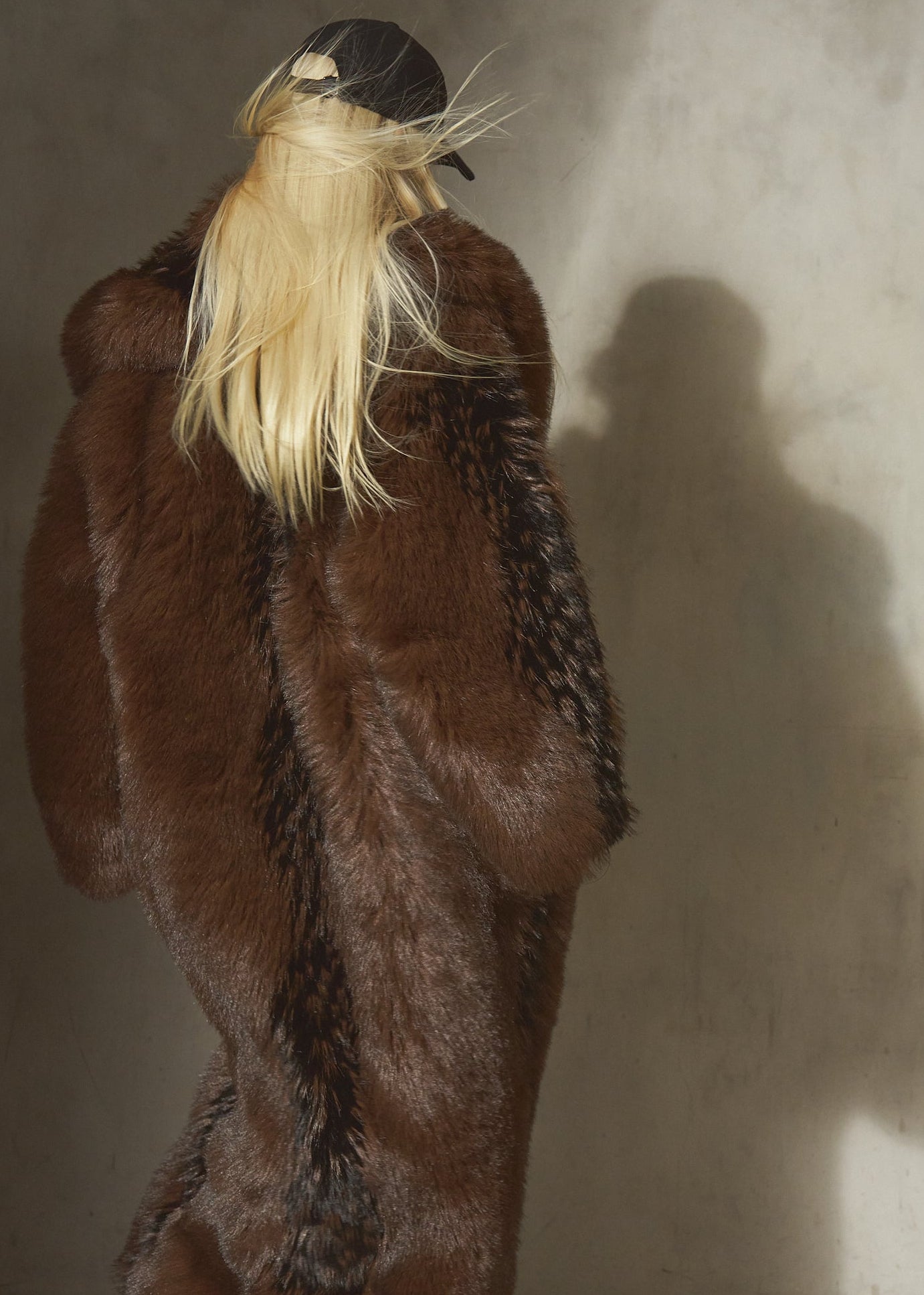 A blonde model viewed from behind wearing a voluminous dark brown and black faux fur floor length coat.