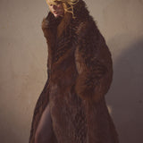Female model wearing a faux-fur brown and black floor length coat against a cement background.