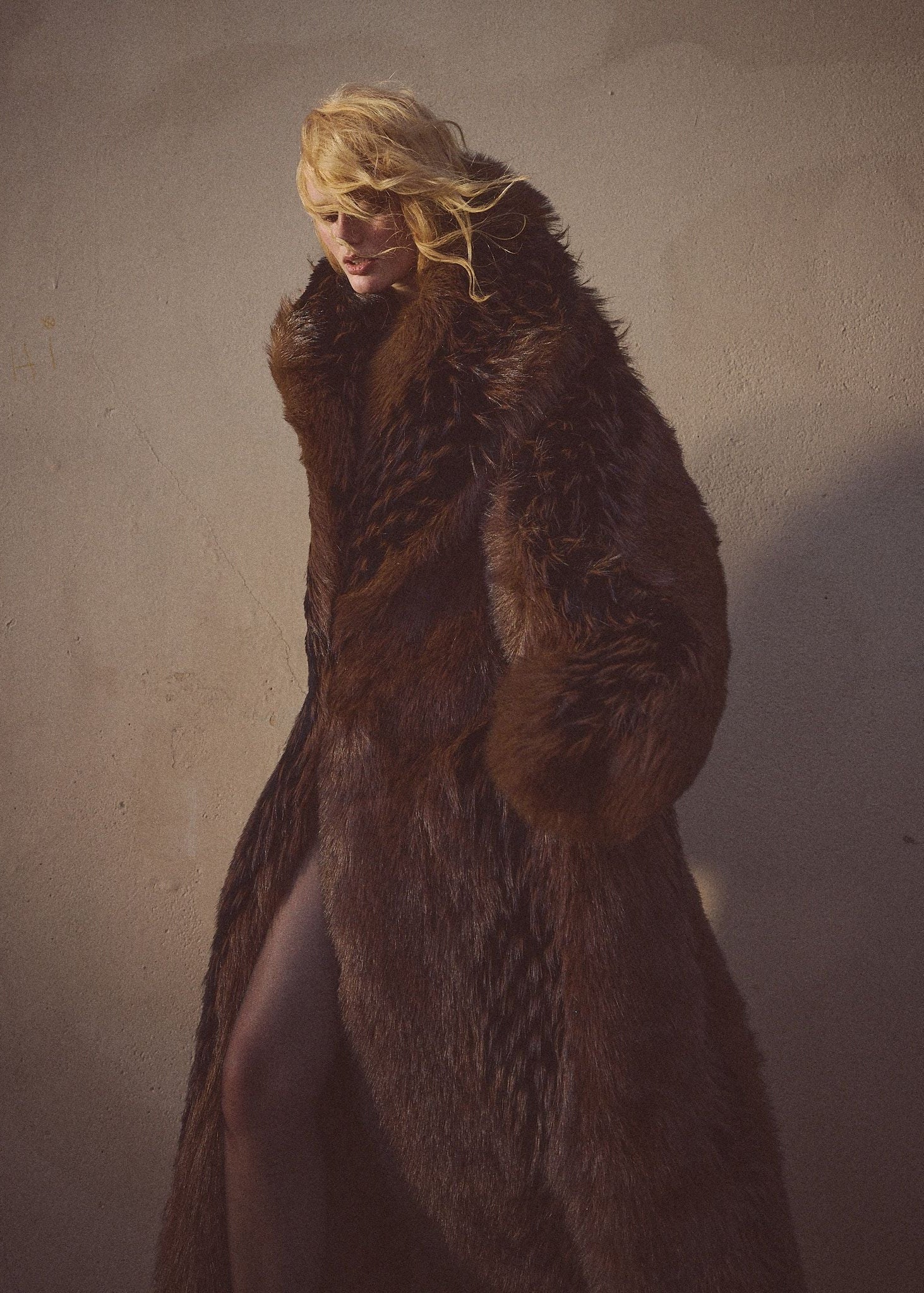 Female model wearing a faux-fur brown and black floor length coat against a cement background.