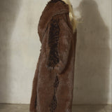 A woman stands in profile wearing a long, luxurious brown and black faux fur coat, styled with a black wool baseball cap, and black heels, set against a textured concrete wall with a strong shadow.