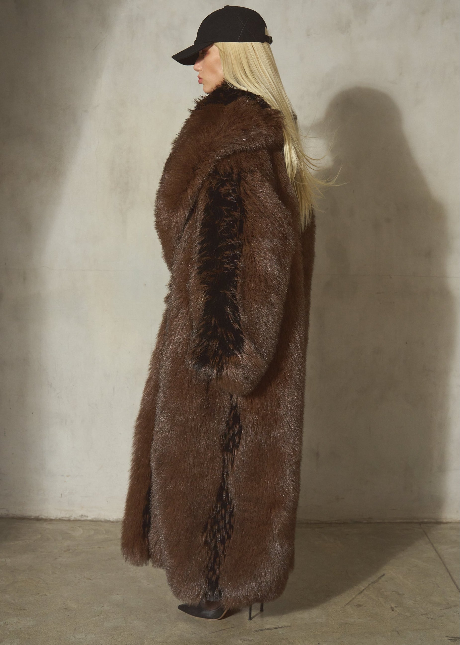 A woman stands in profile wearing a long, luxurious brown and black faux fur coat, styled with a black wool baseball cap, and black heels, set against a textured concrete wall with a strong shadow.