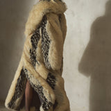 A model wearing a long, luxurious faux-fur coat with beige and black tones, paired with a black wool cap and heels, standing against a textured concrete wall.