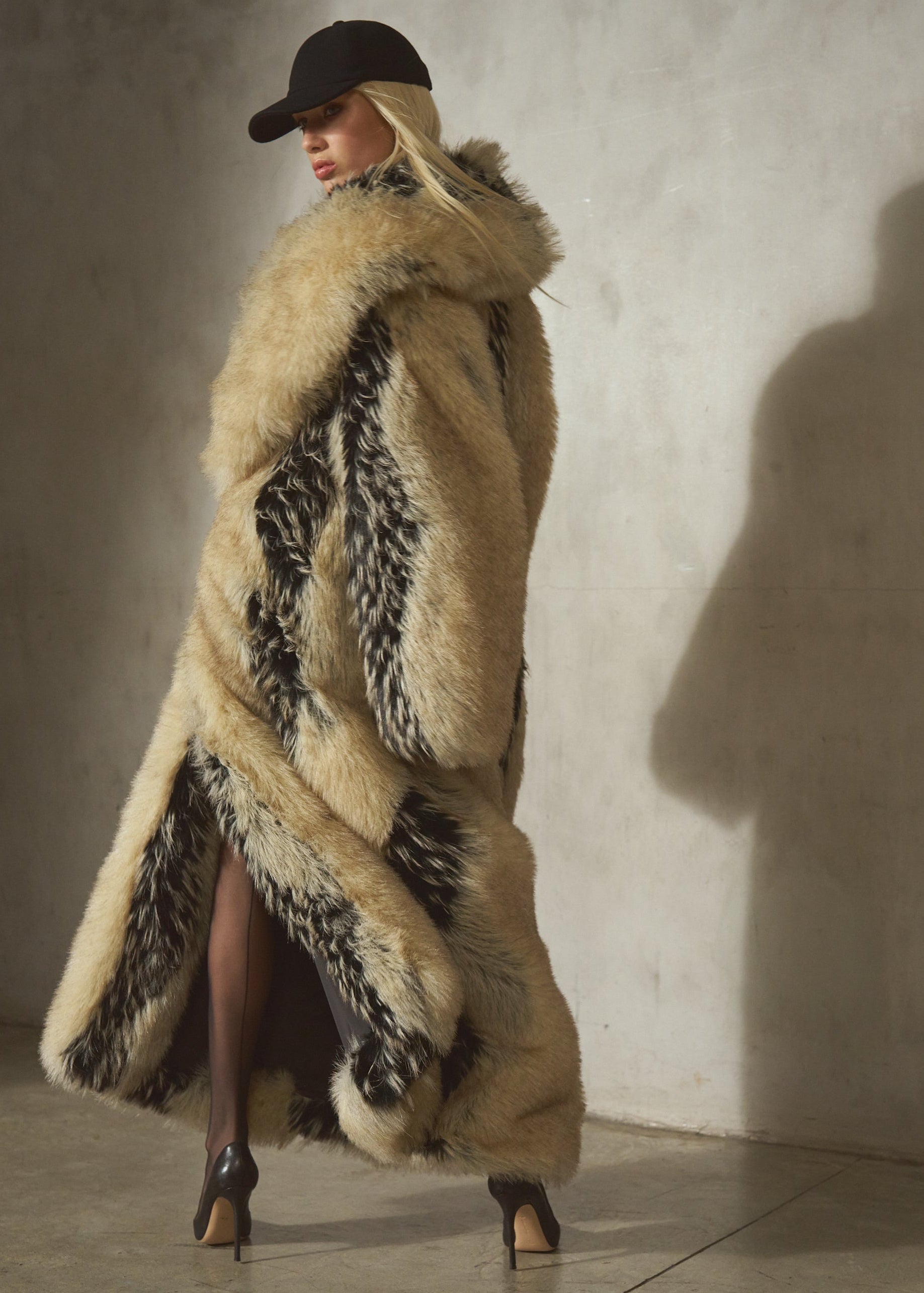 A model wearing a long, luxurious faux-fur coat with beige and black tones, paired with a black wool cap and heels, standing against a textured concrete wall.