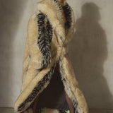 A model wearing a long, luxurious beige and black faux-fur coat with a black cap, standing against a textured concrete wall.