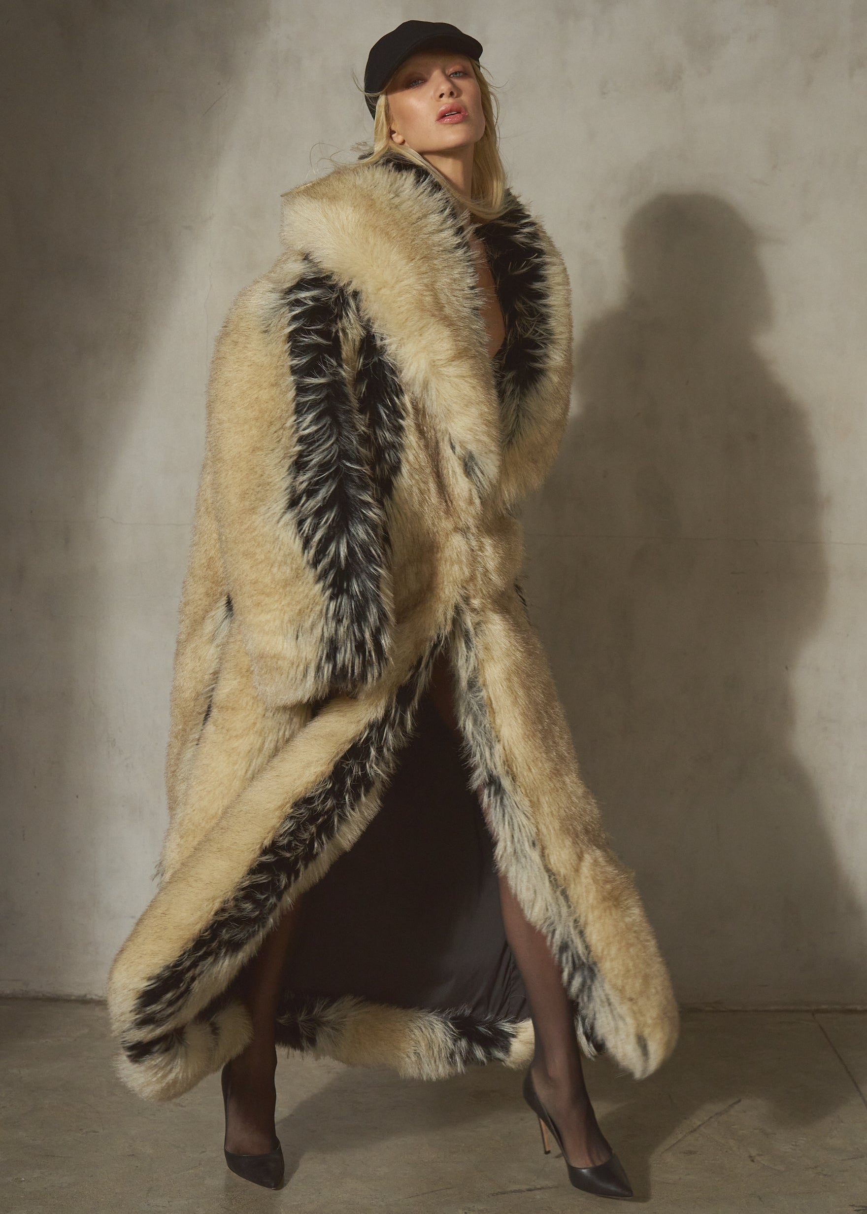 A model wearing a long, luxurious beige and black faux-fur coat with a black cap, standing against a textured concrete wall.