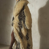 A model wearing a long, voluminous beige and black patterned faux-fur coat, a black wool baseball cap, and black high heels, standing in a studio with a concrete wall and floor.