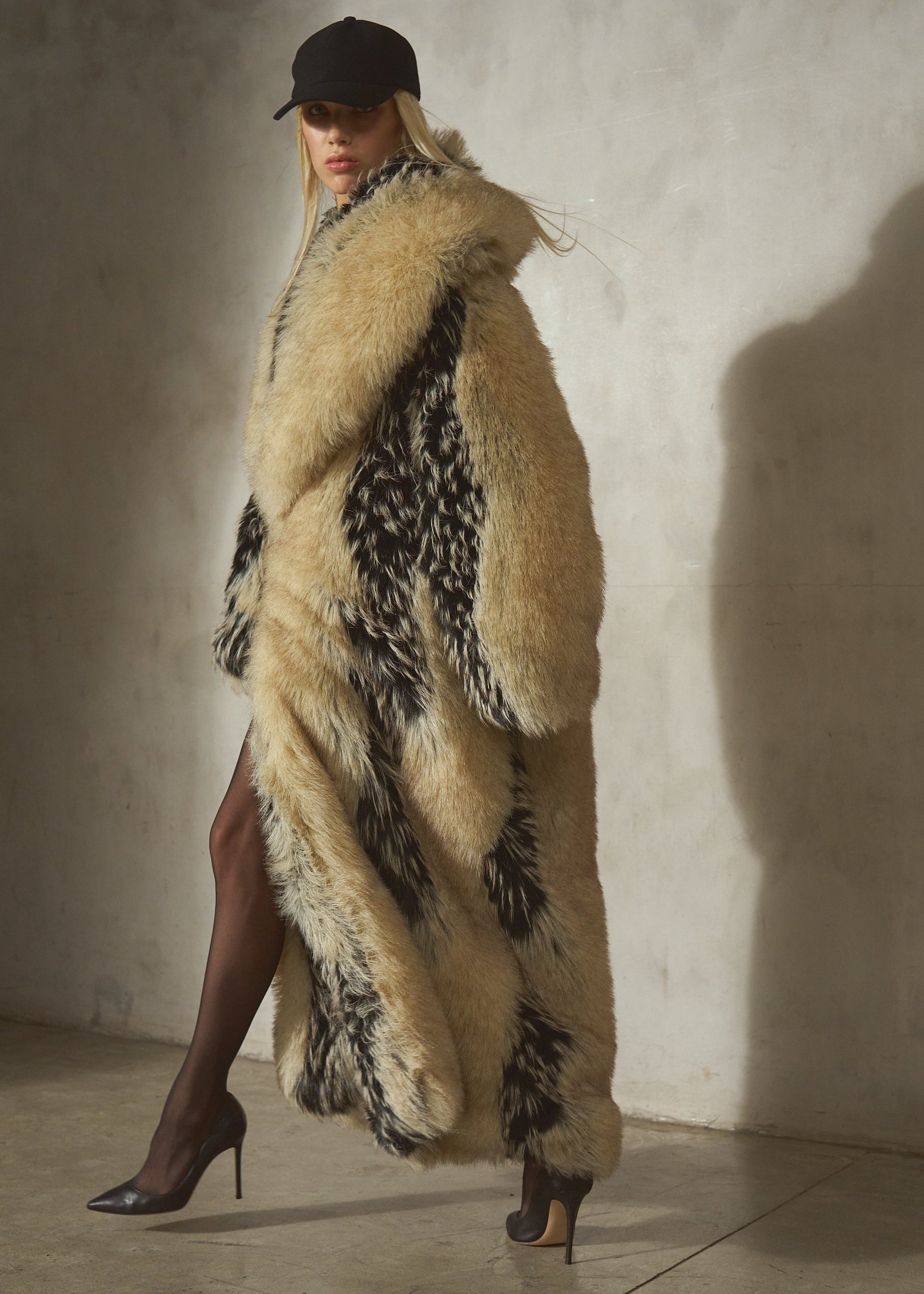A model wearing a long, voluminous beige and black patterned faux-fur coat, a black wool baseball cap, and black high heels, standing in a studio with a concrete wall and floor.