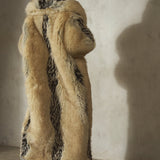 A model viewed from behind wearing a long, luxurious faux-fur coat with beige and black tones, paired with a black wool cap and heels.