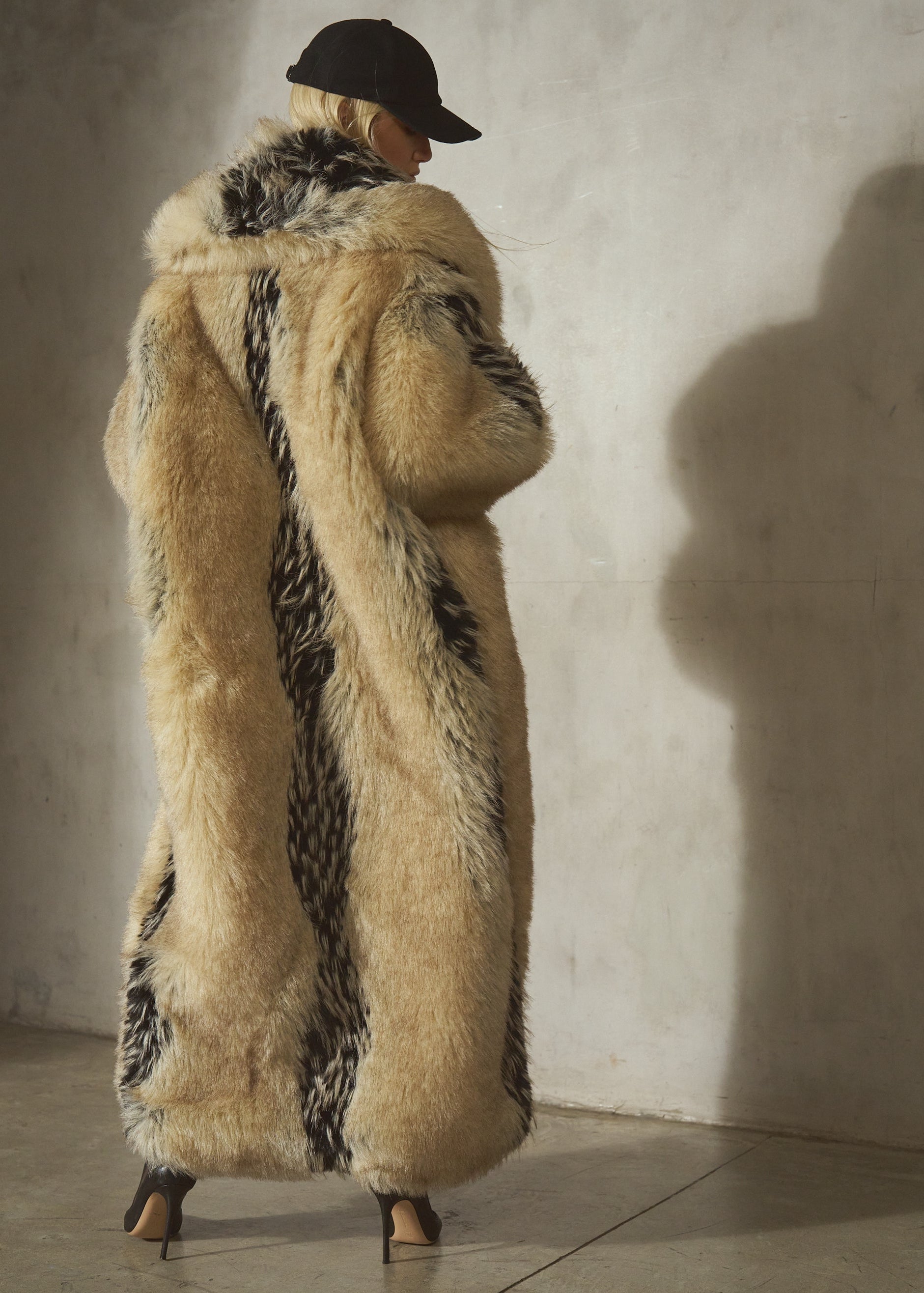 A model viewed from behind wearing a long, luxurious faux-fur coat with beige and black tones, paired with a black wool cap and heels.